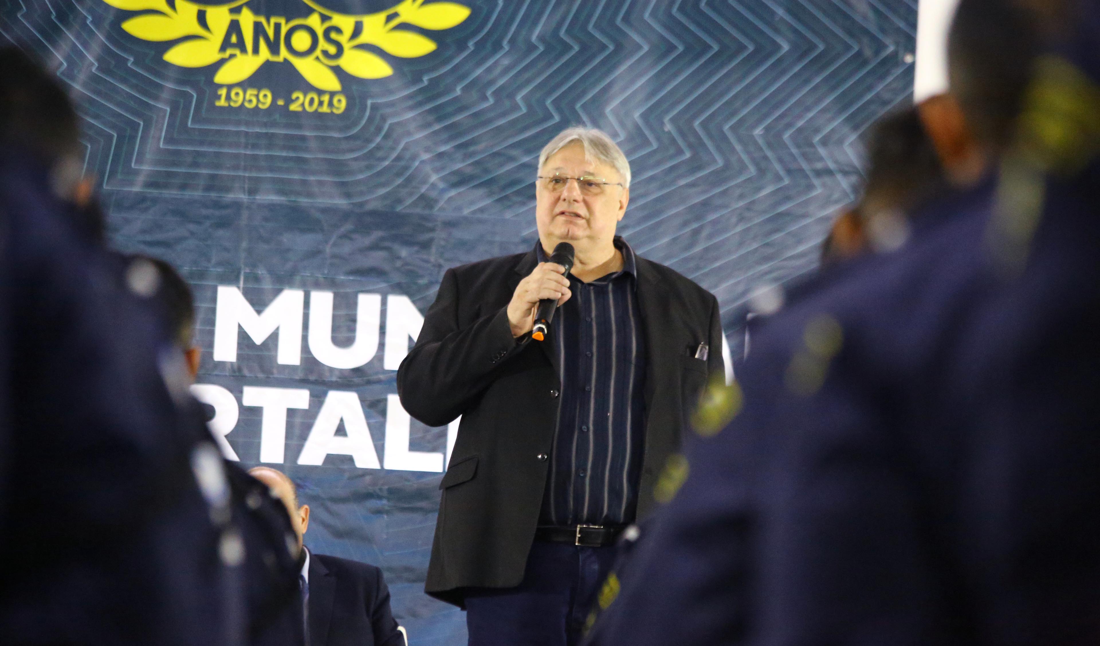 moroni em cima de palco falando ao microfone e guardas municipais em frente em pé